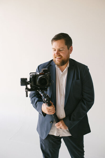 A bearded man holding a camera, positioned against a clean white background.