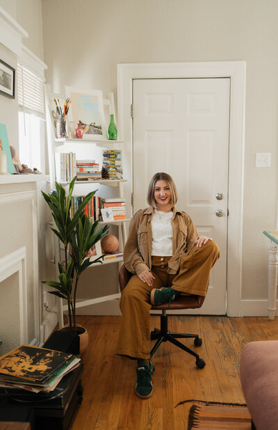 "A woman with a short bob haircut and red lipstick sits casually on a swivel chair in a cozy home office. She wears a tan jacket, white top, mustard-colored pants, and green sneakers. Behind her are white bookshelves filled with books and decorative items, along with a tall green plant."