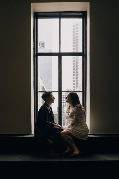 Engaged couple sitting on a window sill, gazing at each other.