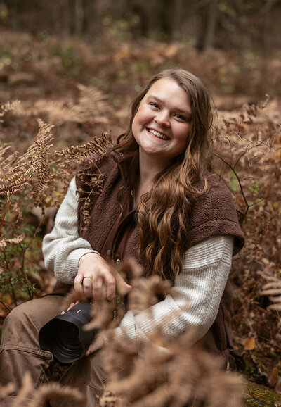 Sydney Breann, a Montana-based elopement photographer, smiles while sitting among autumn ferns holding her camera during a cozy fall portrait session in the forest.