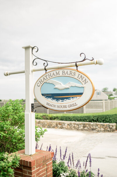 A close-up of the charming white and gold Chatham Bars Inn Beach House Grill sign, framed by summer flowers and a classic Cape Cod stone wall.