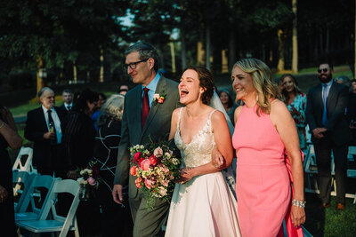Bride laughing while walking down the aisle with mom and dad