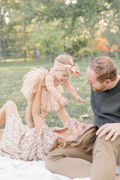a mother lying on a blanket and lifting her baby girl into the air, while the father looks on lovingly at a kansas city park