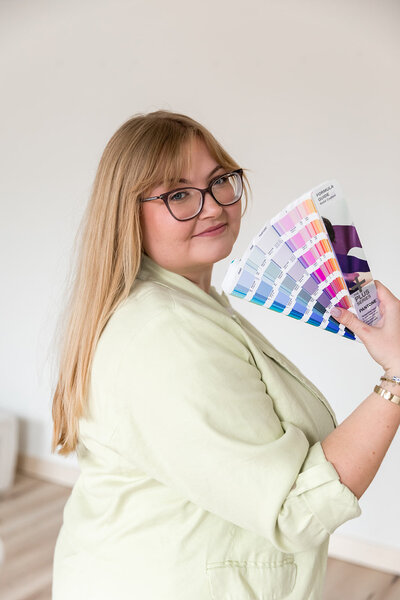 headshot of blonde business owner smiling to the side with glasses. wearing green blazer holding pantone swatches