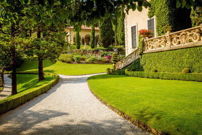 A scenic garden path with neatly trimmed grass and hedges leads to a villa adorned with ivy-covered walls. Flower beds line the area, and a stone balustrade runs along one side. Sunlight filters through the surrounding trees.