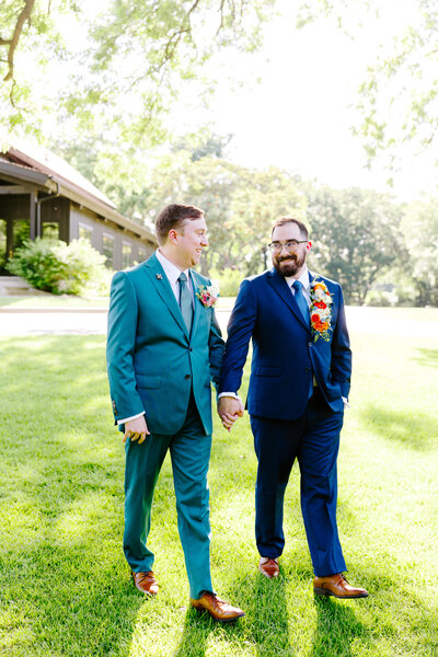 Two grooms walk hand in hand across a sunny lawn during their colorful gay wedding in Oklahoma City, wearing bold suits and floral boutonnieres. Captured by inclusive wedding photographer Bree Smith Photography.

