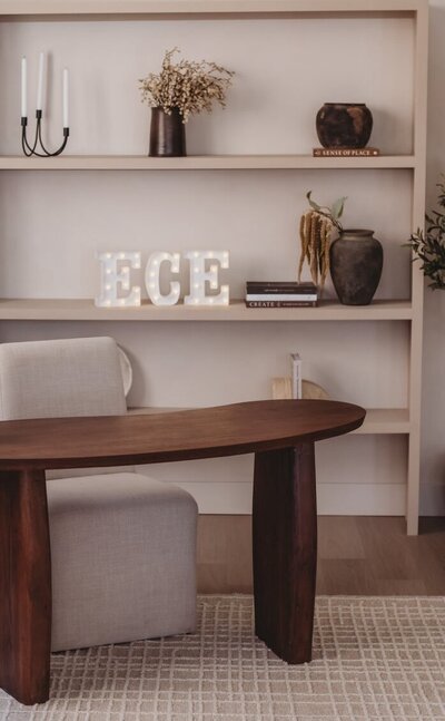 Warm, modern office setup with wooden desk, neutral armchair, and bookshelf featuring decor, books, and illuminated ECE letters.