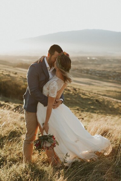 Bride and groom sharing a kiss in a picturesque field with stunning views of Hualalai, capturing a romantic moment from their luxury Hawaii destination wedding.