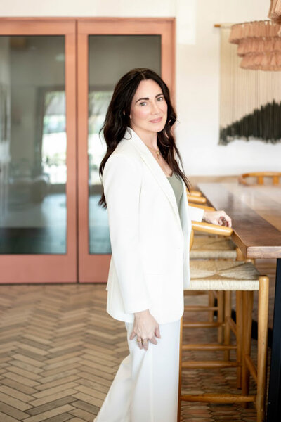 woman with brown hair looking back over her shoulder with hand resting on chair arm.