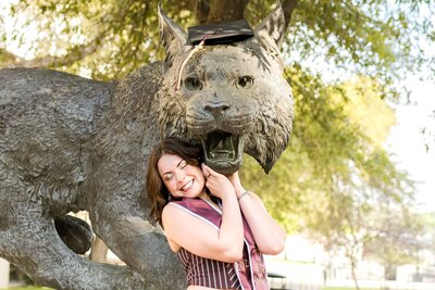 Texas State graduate hugging Bobcat mascot statue and placing graduation cap on statue's head during senior session in San Marcos, Texas.