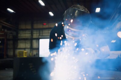 a welder with mask on works over metal, sparks flying everywhere