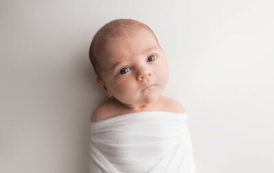 A newborn baby wrapped snugly in a white blanket, gazing wide-eyed at the camera, set against a minimalist white background.