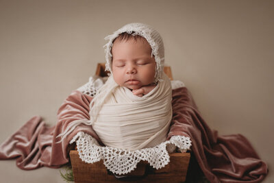 Atlanta, Georgia newborn photographer portrait session. Marietta, Georgia newborn photography session in studio with newborn baby girl wrapped in creme swaddle wearing a cream bonnett and propped up in basket with lace fabrics.