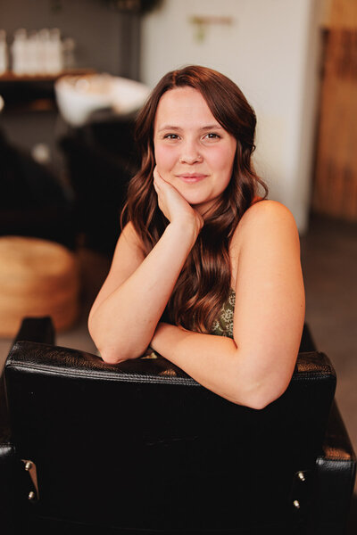 Yasmin a hair stylist in Eugene leans over a chair with her hand on her chin and smiles at the camera.
