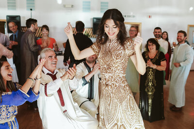 A woman in a gold embroidered dress dances joyfully at a wedding celebration, surrounded by guests in traditional attire who cheer and clap. The lively scene captures the energy of a cultural wedding reception.
