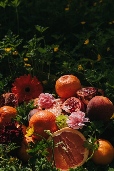 Oranges, pomegranates and flowers lay atop one another outdoors in abundant, lush display.