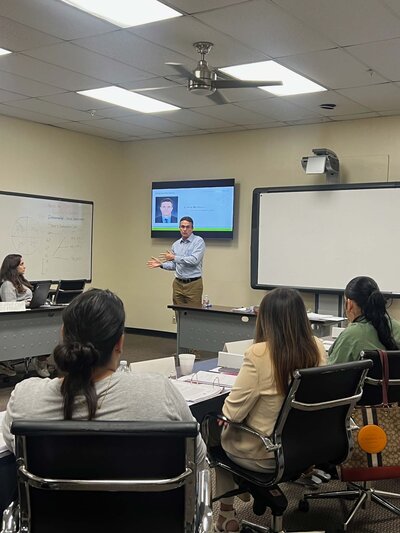 Dallas financial consultants specializing in small businesses attending a conference organized by the Hispanic Chamber of Commerce. The professional is presenting in a classroom setting equipped with a TV.