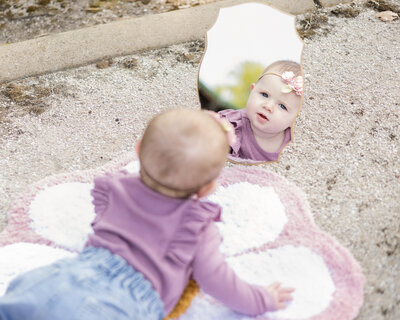 6 month old girl laying on a flower rug on her stomach looking at her reflection in the mirror, wearing a headband with pink flowers, a long sleeve purple shirt and blue jean shorts