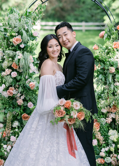 wedding couple pose at vineyard estate next to flower arch in white lace dress and tux