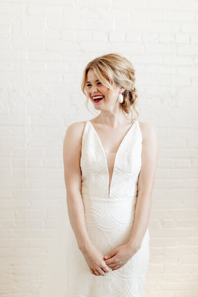 A smiling woman in a white wedding dress with a plunging neckline stands gracefully against a white brick wall. Her wavy blonde hair is styled up, and she wears large white earrings, caught in a candid moment by a Minnesota photographer known for stunning bridal and family portraits.