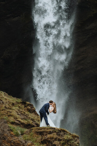 bride and groom are kissing in front of a dramatic waterfall in Iceland during the summer time