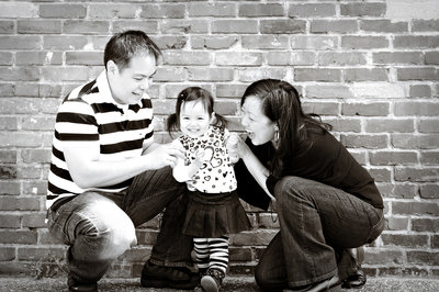 Family of three in front of a brick wall with their little daughter dancing and mom and dad laughing
