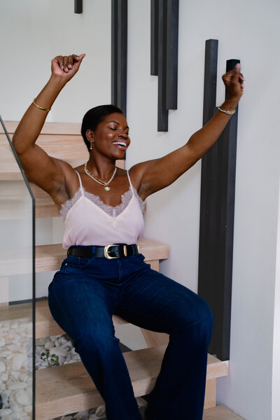 Black career coach for working women raising arms up while sitting on stairs wearing jeans and pink top