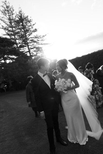 A bride and groom sneak a gaze at each other as the sunset peaks through and their guests are behind them