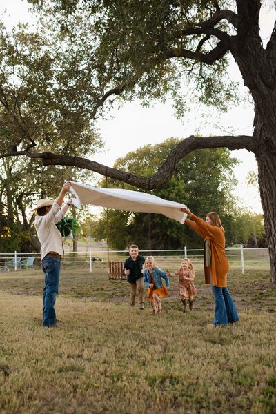A young family plays in the backyard of Liliflora Farmhouse. Mom and dad fluff a blanket while kids run under laughing and smiling. Capture by Shelly Voss