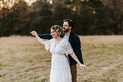 outdoor engagement photos in  little rock ar with man standing behind his fiance as they dance together in a field while the sun sets over the treeline