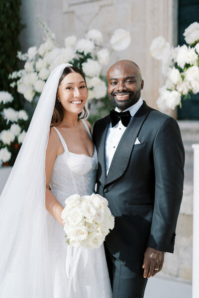 Bride holding white rose bouquet, standing with groom in tuxedo, both smiling at camera after ceremony
