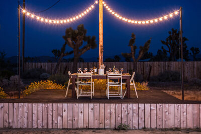 Location branding image Gatos Trail Ranch outdoor dining table set for dinner on stage under with string of lights at dusk
