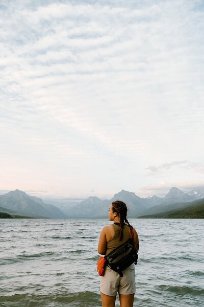 woman standing infront of a huge lake, looking to her left with  huge mountains in the background