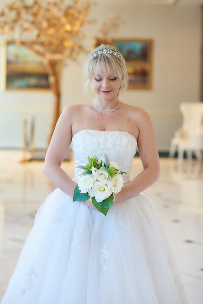 A serene bride with a gentle smile, wearing a sparkling tiara and a pearl necklace, holds a bouquet of white roses. She stands in an elegant indoor setting with soft natural light and a decorative golden tree in the background.