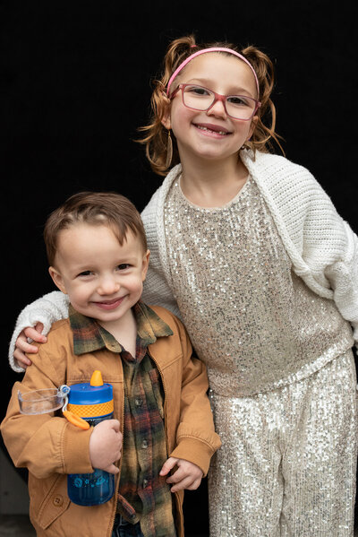 Brother and sister standing in front of a black backdrop, sister with her arm around her younger brother, smiling at the camera for personality portraits