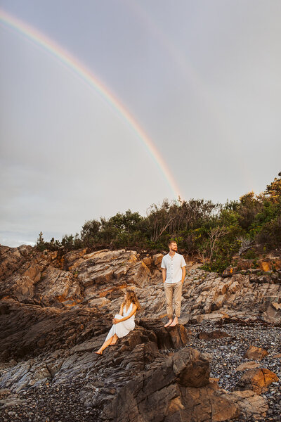 A girl sitting on the rocks looking out over the ocean while the man is standing looking the opposite direction with a rainbow over them.