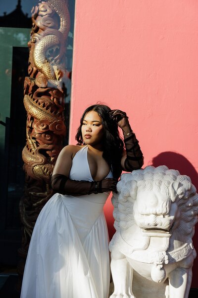 Editorial portrait of a stylish young woman in a white gown and sheer black gloves posing confidently next to a white lion statue in front of a bold pink and red architectural backdrop