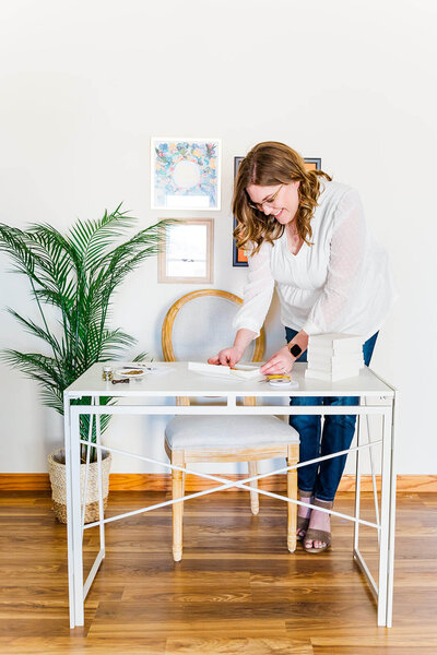 brunette woman standing next to desk looking down at objects