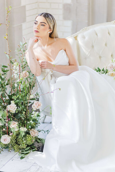 A bride sits on a white couch with roses standing next to her. She has her chin on her hand. Photo taken at Laurel Hall.
