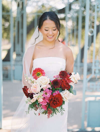 Bride smiling and holding a bold red and blush bouquet in front of a vintage gate, styled for an Austin wedding with timeless charm.
