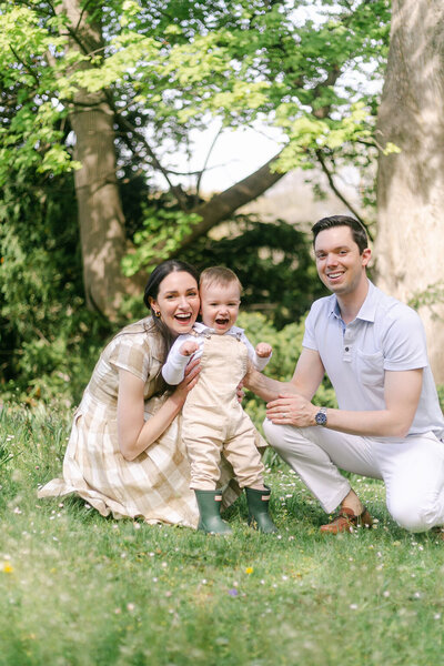 A toddler smiles while playing in the grass with his parents - photo by Rochester NY newborn photography service provider Emi Rose Studio.