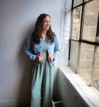 Woman with dark hair in green jumpsuit and jean jacket looking out window
