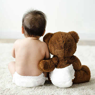 A child wearing a diaper sitting away from camera and a teddy bear with a diaper