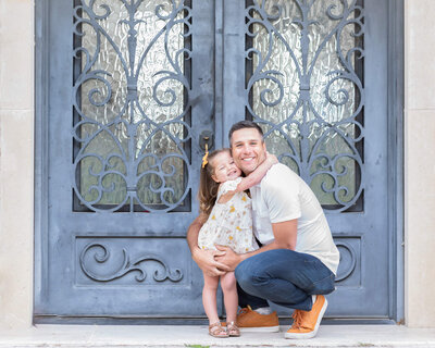 Father crouched down for toddler daughter wearing pretty dress to give him a neck hug in front of a door in Adriatica McKinney TX