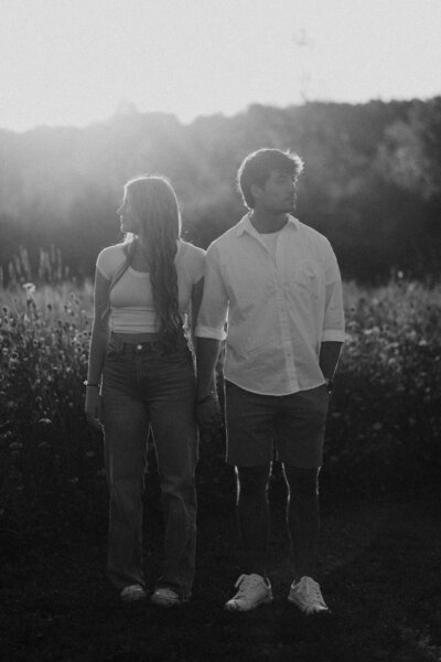 A couple stands in a sunlit field holding hands, gazing apart. The black-and-white image creates a nostalgic, serene atmosphere.