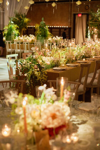 A close-up of an elegant table setting featuring three ornate glasses, one empty and two with champagne. A lit candle in a brass holder stands on the right, with soft-focus greenery and more candles in the background, creating a warm and festive ambiance.