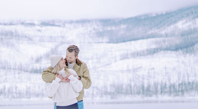 Engaged couple embracing in a snowy mountain landscape, dressed in winter outfits and sharing a quiet, romantic moment, captured by Joseph Mayden during a seasonal engagement session.