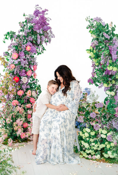 A pregnant woman in a flowy white and blue floral dress sits on a chair, smiling as a young boy hugs her belly, surrounded by lush floral arches of purple, pink, and green blooms.