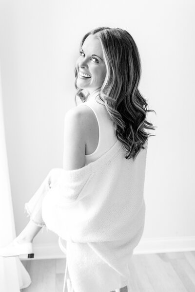 A black and white portrait of a photographer in a white off the shoulder sweater smiling over her shoulder in a Kansas City photography studio. 