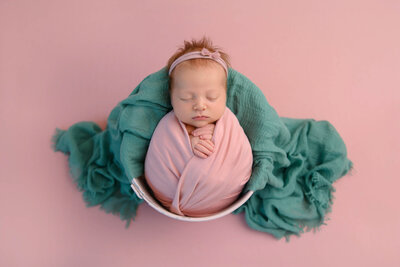 red-haired newborn baby girl wrapped in pink in a pink bucket with a green blanket for her Burlington, Ontario newborn photography session.